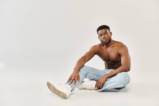 Young shirtless African American man sitting on the ground in a studio with a grey background. - Photo, Image