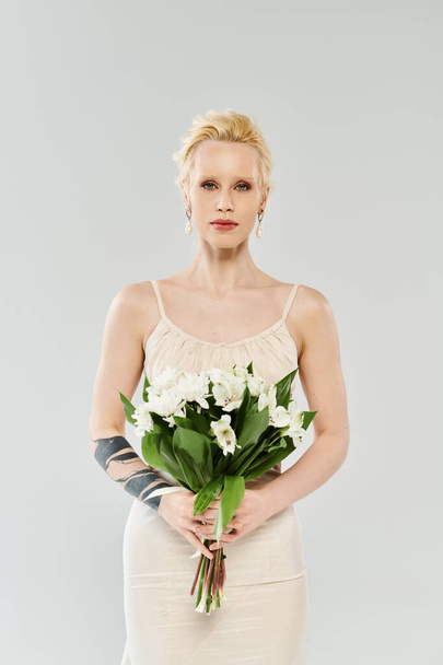Novia rubia etérea en vestido blanco, con elegancia sosteniendo vibrante ramo de flores sobre un fondo gris. - Foto, imagen