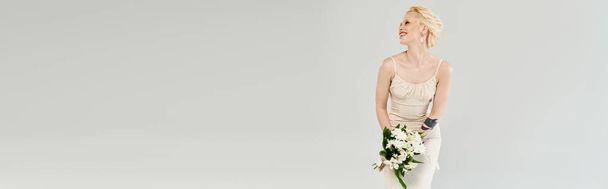 Egy elbűvölő szőke menyasszony áll kecsesen fehér ruhában, finoman tartva egy buja csokor élénk virágot.. - Fotó, kép