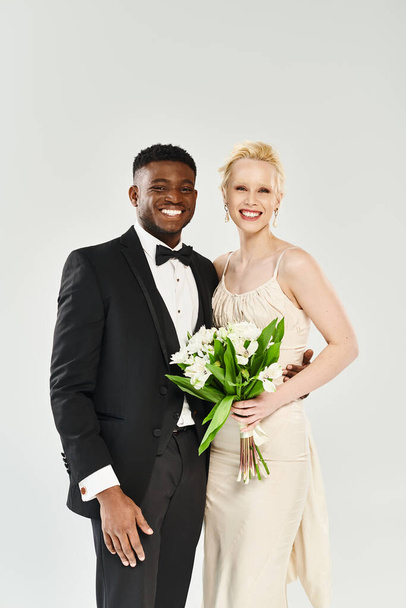 A beautiful blonde bride in a wedding dress and an African American groom in a tuxedo stand elegantly on a grey studio background. - Photo, Image