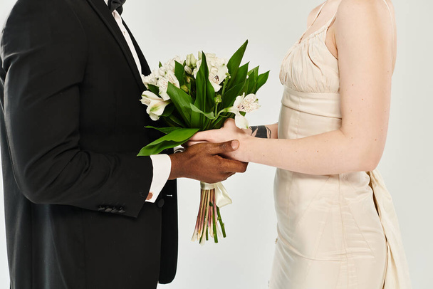 Ein afroamerikanischer Bräutigam im Smoking hält einen Blumenstrauß neben einer schönen blonden Braut in ihrem Hochzeitskleid. - Foto, Bild
