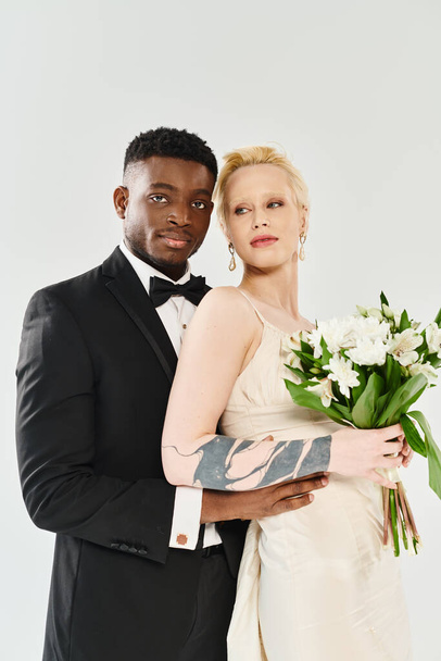 A beautiful blonde bride in a wedding dress and an African American groom in a tuxedo holding a bouquet on a grey background. - Photo, Image