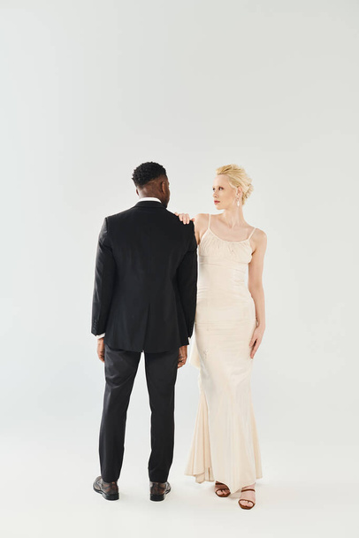 Une belle mariée blonde dans une robe de mariée et un marié afro-américain se tiennent côte à côte dans un studio sur un fond gris. - Photo, image