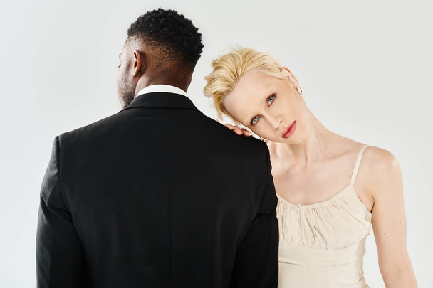 Eine schöne blonde Braut im Brautkleid und ein afroamerikanischer Bräutigam stehen nebeneinander in einem Studio vor grauem Hintergrund.. - Foto, Bild