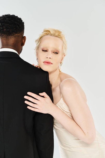 A beautiful blonde bride in a white dress and an African American groom in a tuxedo stand together on a grey studio background. - Photo, Image
