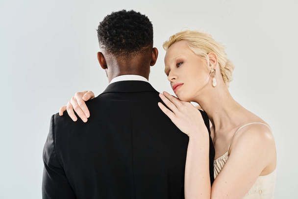 A beautiful blonde bride in a wedding dress and an African American groom embracing each other passionately in a studio on a grey background. - Photo, Image