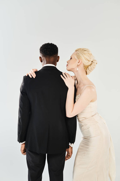 Piękna blond panna młoda w sukni ślubnej stoi obok Afroamerykańskiego pana młodego w studio na szarym tle. - Zdjęcie, obraz