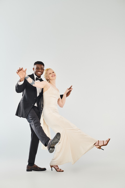 Eine schöne blonde Braut im Brautkleid und ein afroamerikanischer Bräutigam tanzen anmutig zusammen in einem Studio auf grauem Hintergrund. - Foto, Bild