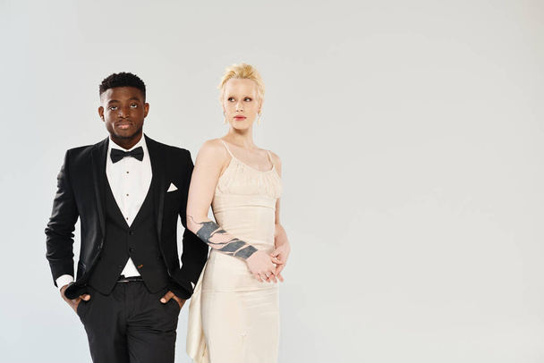 An African American man in a tuxedo stands next to a beautiful blonde woman in a white dress in a studio against a grey background. - Photo, Image