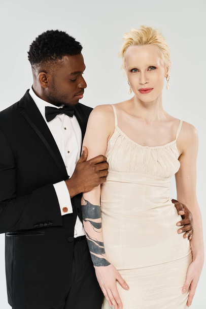 Африканський наречений і красива блондинка наречена, одягнена в смокінг і сукню, витончено стоїть в студії на сірому фоні. - Фото, зображення