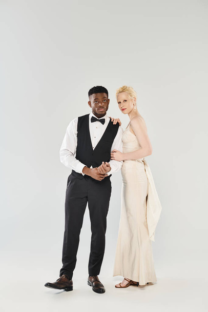 An African American groom in a tuxedo and a beautiful blonde bride in a flowing wedding dress pose elegantly in a studio setting. - Фото, изображение