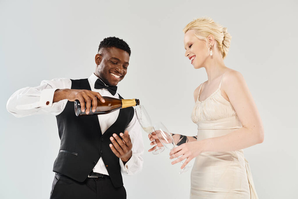 Чоловік в смокінгу вливає шампанське в жіночу руку, оскільки вони святкують в студії з красивою блондинкою нареченої у весільній сукні та афроамериканським нареченим на сірому фоні. - Фото, зображення