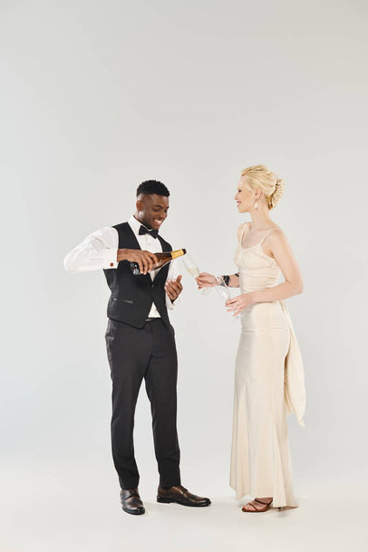 Een mooie blonde bruid in een trouwjurk en een Afro-Amerikaanse bruidegom die naast elkaar staan in een studio op een grijze achtergrond. - Foto, afbeelding