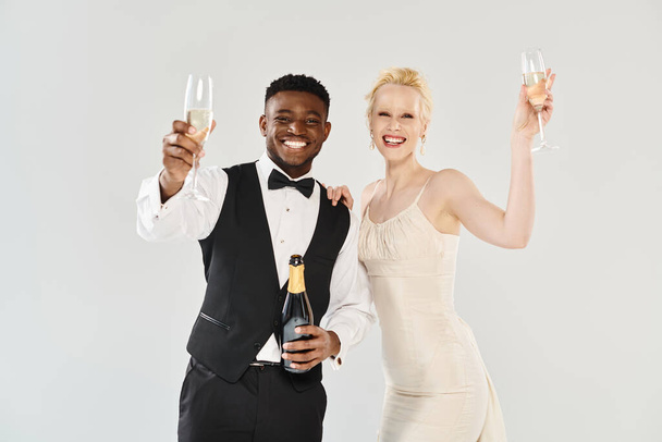 Eine schöne blonde Braut im Brautkleid und ein afroamerikanischer Bräutigam mit Champagnergläsern in einem Studio vor grauem Hintergrund. - Foto, Bild