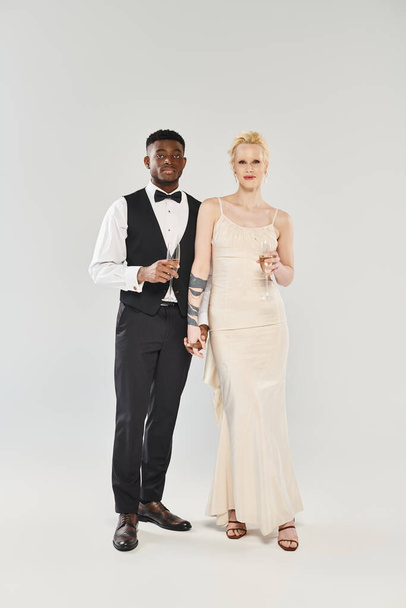 Uma noiva loira bonita em um vestido de noiva e um noivo afro-americano em um smoking ficar juntos em um estúdio contra um fundo cinza. - Foto, Imagem