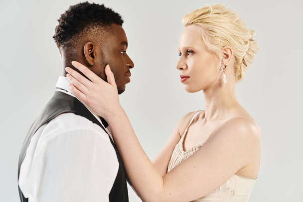 Piękna blond panna młoda w sukni ślubnej i afro-amerykański pan młody stojący razem w studio na szarym tle. - Zdjęcie, obraz
