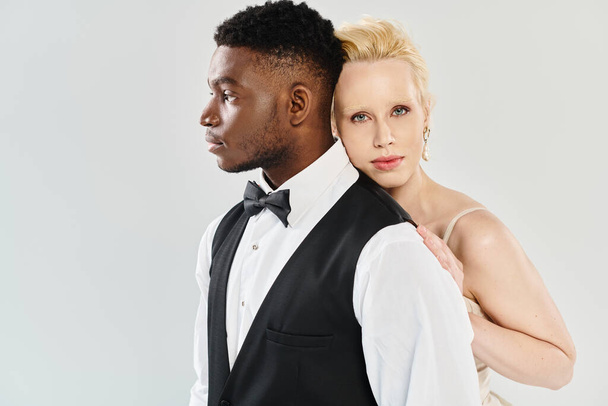 A beautiful blonde bride in a wedding dress standing next to an African American groom in a tuxedo against a grey studio backdrop. - Photo, Image