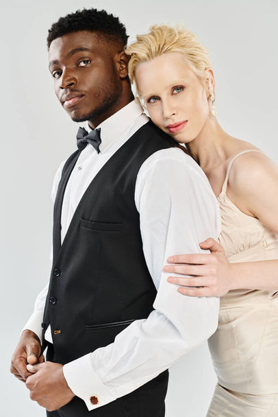 Uma noiva loira bonita em um vestido de noiva e um noivo afro-americano em um smoking ficar em um estúdio contra um fundo cinza. - Foto, Imagem