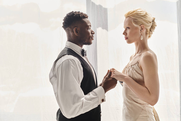 Una hermosa novia rubia con un vestido de novia y un novio afroamericano de pie juntos en un estudio, sobre un fondo gris. - Foto, Imagen