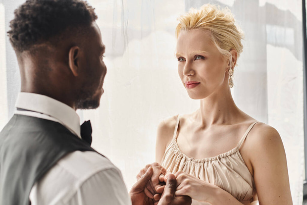 Eine schöne blonde Braut im Brautkleid und ein afroamerikanischer Bräutigam stehen nebeneinander in einem Studio vor grauem Hintergrund.. - Foto, Bild