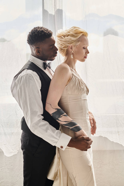 Красива блондинка в весільній сукні і афроамериканський наречений стоїть поруч один з одним в студії на сірому фоні. - Фото, зображення