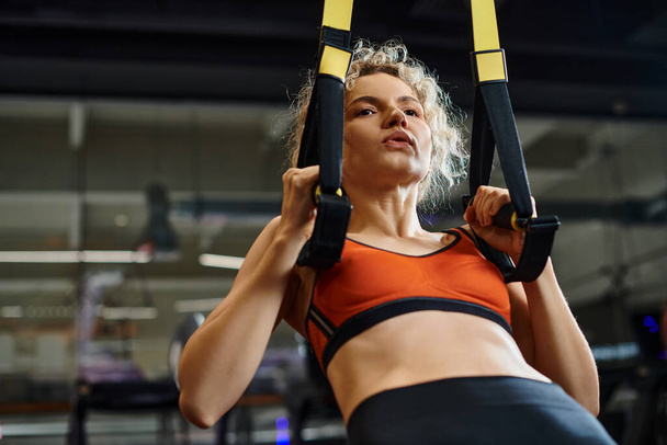 good looking blonde woman in comfortable sportwear exercising with pull ups equipment while in gym - Photo, Image