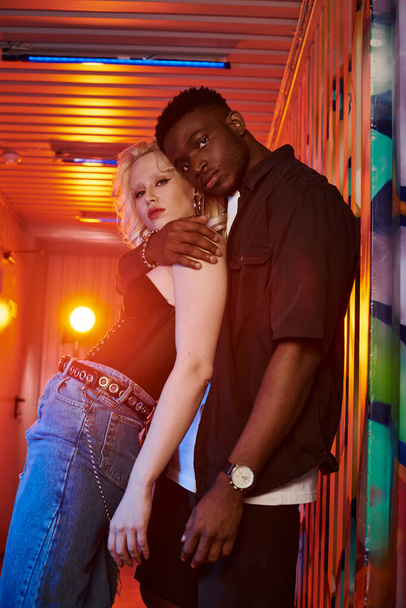 Una donna bionda e un uomo afroamericano stanno fianco a fianco su una strada urbana con graffiti colorati sulle pareti. - Foto, immagini
