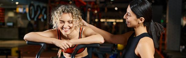 entraîneur féminin joyeux aidant son beau client à utiliser cross trainer dans la salle de gym, bouteille d'eau, bannière - Photo, image