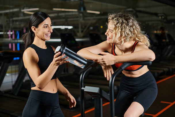attrayant femme énergique en utilisant moderne cross trainer tandis que dans la salle de gym à côté de son entraîneur féminin athlétique - Photo, image