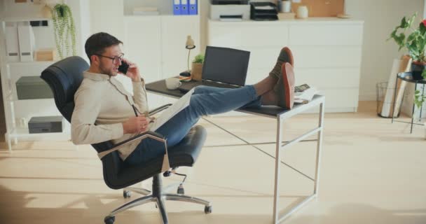 Профессионал мужского бизнеса, разговаривающий по смартфону и изучающий документы, сидя в расслабленной позе на стуле в офисе - Кадры, видео