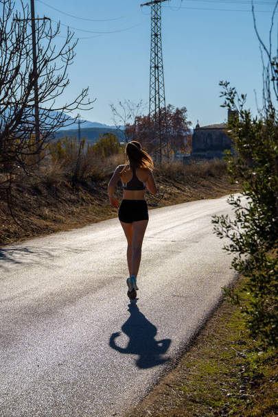 Sylwetka młodej biegaczki podświetlona światłem słonecznym o świcie, ćwicząca wyścig po wybrukowanej ścieżce w parku naturalnym, rzucająca cień na asfalt. - Zdjęcie, obraz