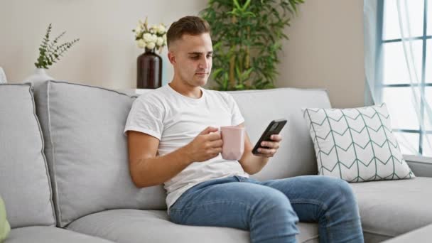 Mladý muž pomocí smartphone na pohovce drží růžový hrnek v útulném obývacím pokoji - Záběry, video