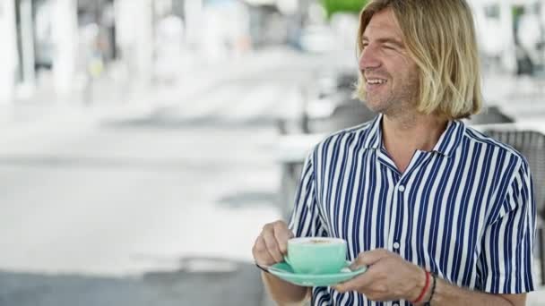 Een stijlvolle man met lang haar geniet van een kopje cappuccino op een terras van de stad, belichaamt stedelijke recreatie en mode. - Video
