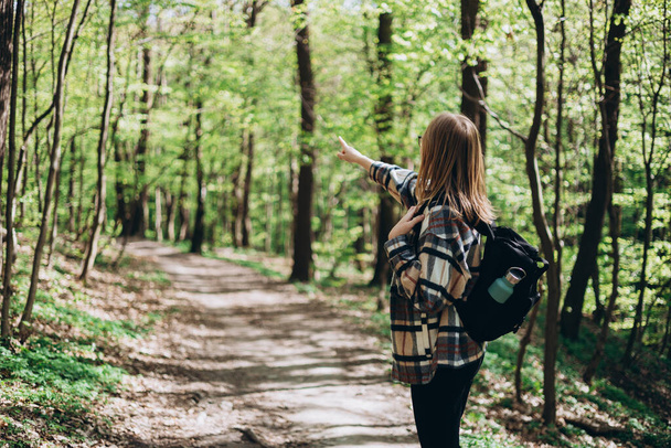 Rubia chica de 30 años en gafas apuntando con el dedo sobre el fondo del bosque. Joven hembra con mochila senderismo en la naturaleza. Hermosa mujer caminando en ropa deportiva. Concepto de estilo activo - Foto, Imagen