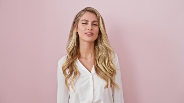 Vrolijke, sexy blonde vrouw die een liefdeskus naar de camera blaast. staand in roze, geïsoleerde achtergrond met een shirt aan, die schattige energie uitdrukt. - Video