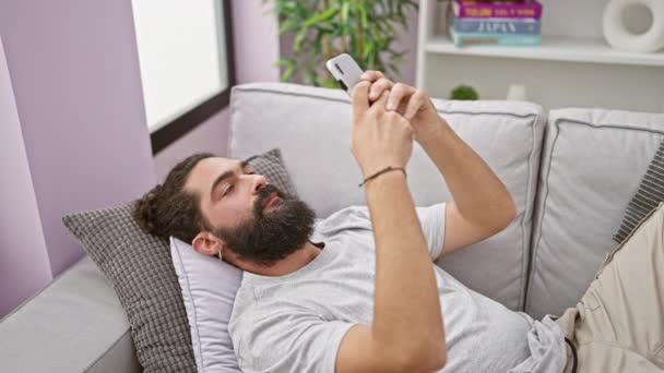 Handsome hispanic man lounging on a sofa with a smartphone in a cozy modern living room, exemplifying relaxed home lifestyle. - Footage, Video
