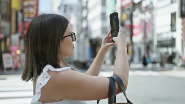 Egy boldog, gyönyörű spanyol nő szemüvegben tokyo városi tájképét örökíti meg okostelefonnal, elmerülve Japán építészeti szépségében. - Felvétel, videó