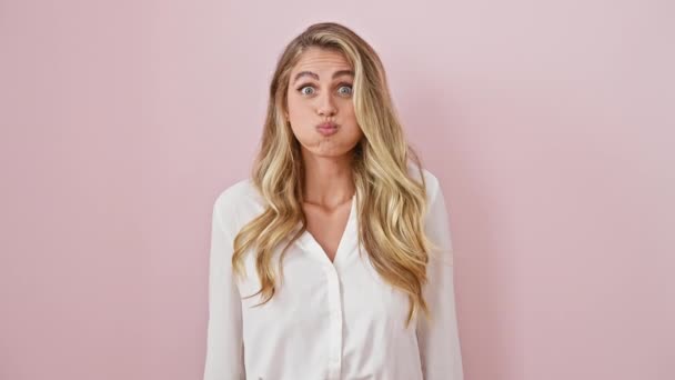 Lustiges Porträt einer jungen blonden Frau, die vor rosa Hintergrund die Wangen aufbläst, steht und einen verrückten, lustigen Gesichtsausdruck bricht - Filmmaterial, Video