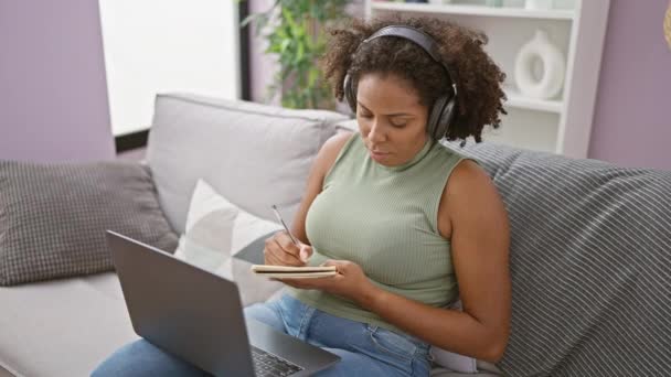 Afrikkalainen amerikkalainen nainen kirjoittaa muistiinpanoja sohvalla kannettavan tietokoneen ja kuulokkeet - Materiaali, video
