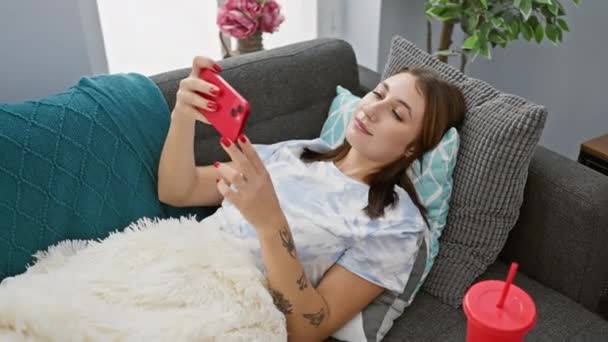 Μια χαλαρή γυναίκα ξαπλώνει σε έναν καναπέ, απορροφημένη στο smartphone της, σε ένα σπιτικό καθιστικό με φωτεινές προφορές. - Πλάνα, βίντεο