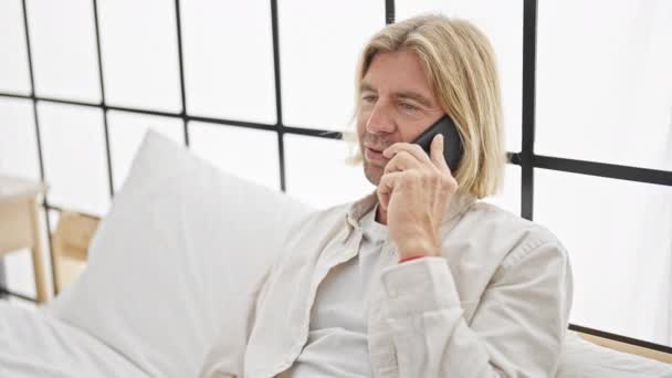 Yakışıklı, uzun saçlı bir adam. Parlak, modern bir yatak odasında bir yatakta dinlenirken telefonla konuşuyor. - Video, Çekim