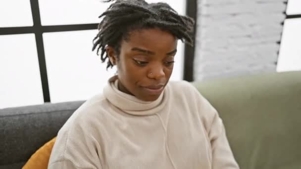 Überraschung in den Augen, junge schwarze Frau mit Dreadlocks, selbstbewusst zu Hause auf einem gemütlichen Sofa sitzend, humorvoll mit positiver, fröhlicher Miene durch die "ok" -Geste schauend.. - Filmmaterial, Video