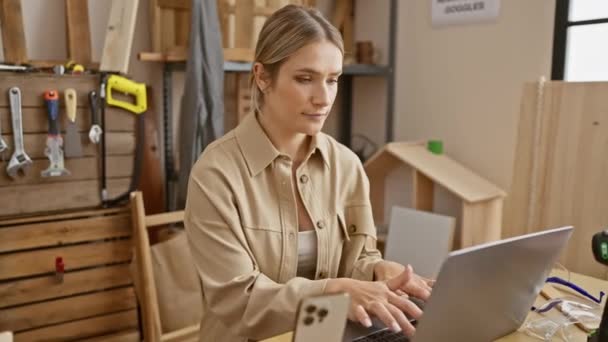 Jonge, vastberaden blonde vrouw navigeert vol vertrouwen door laptop in timmerwerkplaats, met een serieuze expressie en denkhouding - Video