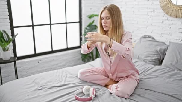 Een jonge vrouw in roze pyjama analyseert een thermometer terwijl ze op een bed zit, met koptelefoon en een moderne interieur slaapkamer achtergrond. - Video