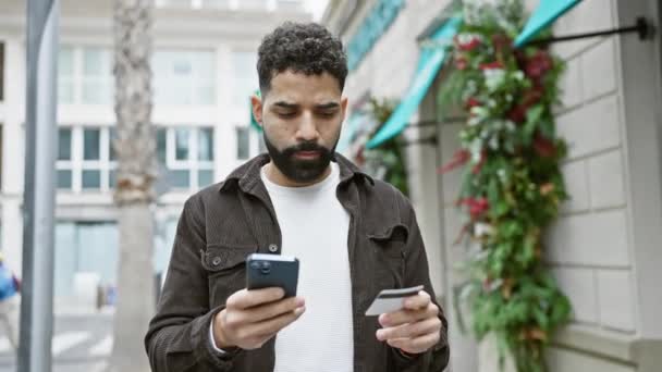 Przystojny Hiszpan z brodą przy użyciu smartfona i karty kredytowej na ulicy - Materiał filmowy, wideo