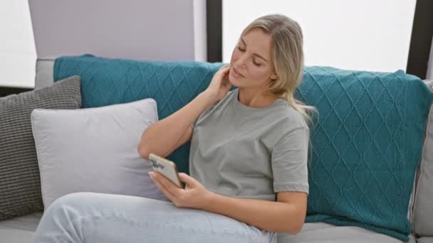 Blonde Frau leidet unter Nackenschmerzen, während sie ihr Smartphone in einem modernen Wohnzimmer benutzt. - Filmmaterial, Video