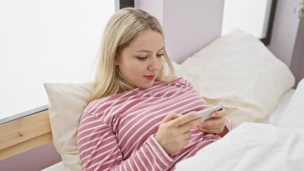 若い女性はベッドで彼女のスマートフォンを使用して楽しんで,親密で居心地の良い,快適な屋内設定を示しています. - 映像、動画