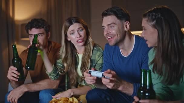 笑顔の仲間が週末のホームハングアウトでボトルをクローズアップ. ハッピーグループの友人が夜遅くに一緒にビールを飲んで座っている. 夜にビデオゲームを楽しむリラックスしたゲーマーが平らに集まる - 映像、動画
