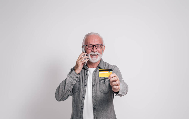 Sorrindo idoso gerente do sexo masculino falando por telefone celular e mostrando cartão de crédito no fundo branco - Foto, Imagem