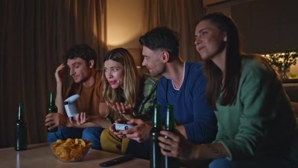 Дружні геймери грають у відеоігри ввечері вдома. Усміхнені молоді, які використовують консолі для відеоігор, сидять у вітальні разом. Щасливі безтурботні друзі насолоджуються розвагами у вихідні в квартирі. - Кадри, відео